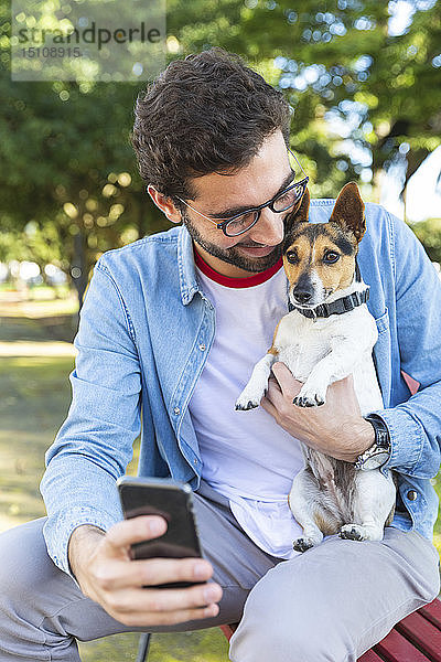 Junger Mann sitzt mit seinem Hund auf der Parkbank und nimmt sich mit seinem Smartphone ein