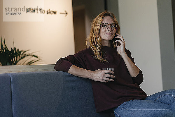Lächelnde junge Frau sitzt auf der Couch und telefoniert mit dem Handy