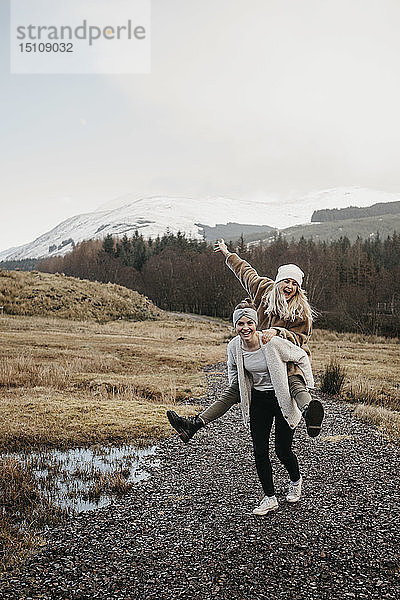 Grossbritannien  Schottland  glückliche junge Frau trägt Freundin huckepack in ländlicher Landschaft