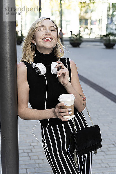 Porträt einer glücklichen blonden Frau mit Kaffee zum Mitnehmen  Umhängetasche und Kopfhörer