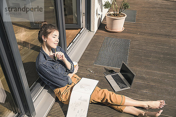 Junge Frau mit geschlossenen Augen sitzt zu Hause auf der Terrasse mit Laptop und Buch