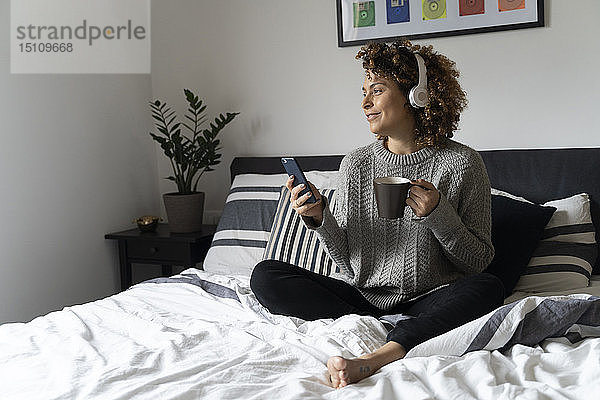 Frau sitzt im Bett  trinkt Kaffee  benutzt Smartphone und Kopfhörer