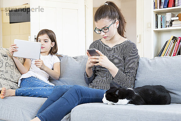 Zwei Schwestern sitzen zu Hause auf der Couch und benutzen elektronische Geräte