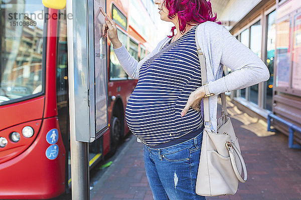 Schwangere Frau an Bushaltestelle mit Fahrplankontrolle