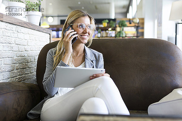 Junge Frau telefoniert mit ihrem Handy  während sie ihr digitales Tablet in einem Café benutzt