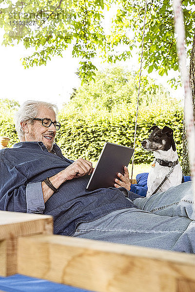 Älterer Mann entspannt sich auf einem Schaukelbett in seinem Garten und benutzt ein digitales Tablet