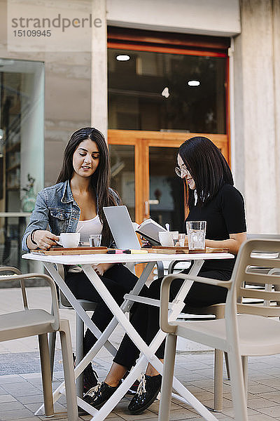 Zwei Freunde sitzen mit Buch und Laptop in einem Straßencafé zusammen