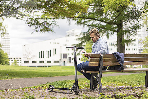 Geschäftsmann mit E-Scooter sitzt auf einer Bank im Stadtpark und benutzt ein Smartphone  Essen  Deutschland