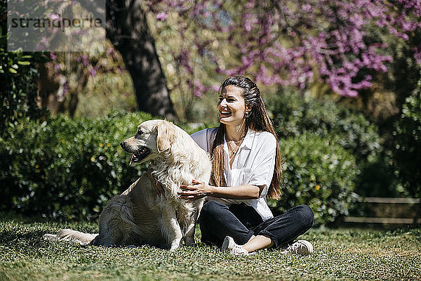 Glückliche Frau sitzt mit ihrem Labrador Retriever auf einer Wiese im Stadtpark und genießt das Sonnenlicht