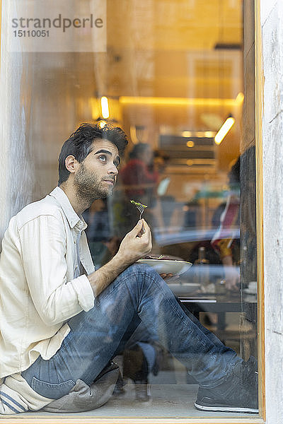 Mann isst einen Salat und sitzt in einem Restaurant am Fenster und schaut hinaus