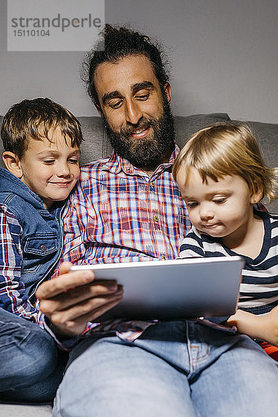 Porträt eines Vaters  der mit seinen Kindern auf der Couch sitzt und sich Filme auf einem digitalen Tablet anschaut