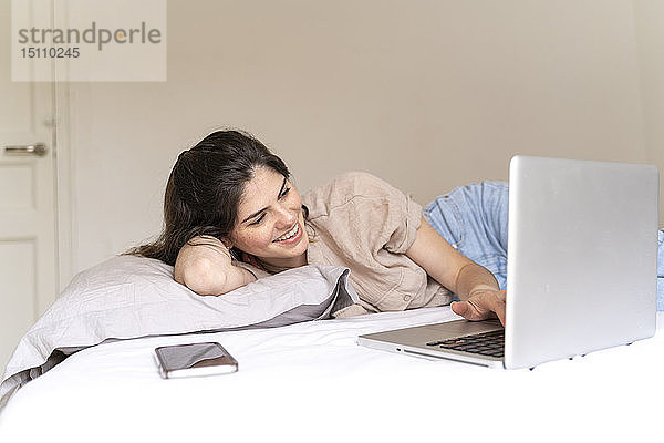Glückliche junge Frau liegt mit Laptop auf dem Bett