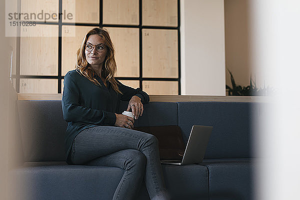 Geschäftsfrau sitzt auf der Couch mit Laptop und Kaffee zum Mitnehmen