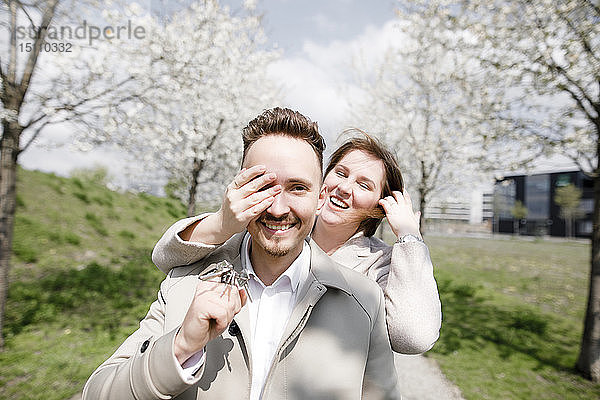 Glücklicher junger Mann hält Riegelschlüssel für neues Haus in der Hand  während die Frau ihm die Augen zudeckt