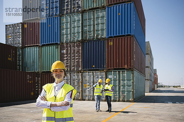 Porträt einer selbstbewussten Arbeitnehmerin vor Kollegen und Frachtcontainern auf einem Industriegelände