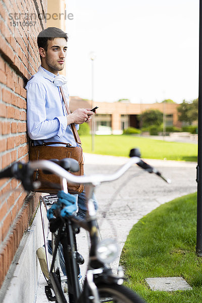 Junger Mann mit Fahrrad und Smartphone  Kopfhörer um den Hals in der Stadt