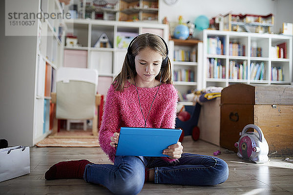 Junges Mädchen  das ein digitales Tablett mit Kopfhörern benutzt und auf dem Boden sitzt