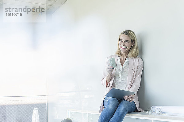 Glückliche Geschäftsfrau sitzt im Büro auf einem Regal mit Tablett