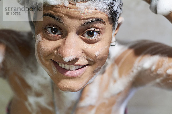 Lächelnde junge Frau  die mit Seife am Körper duscht und in die Kamera schaut
