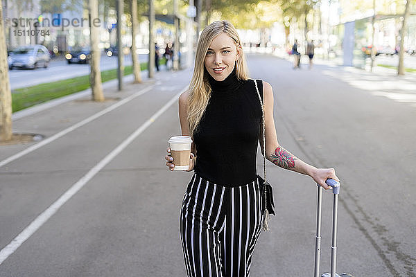 Porträt einer lächelnden blonden Frau mit Kaffee zum Mitnehmen und Rollgepäck