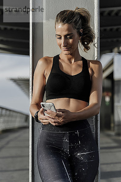Sportliche Frau mit Kopfhörern auf der Brücke stehend  Musik hörend  mit Smartphone