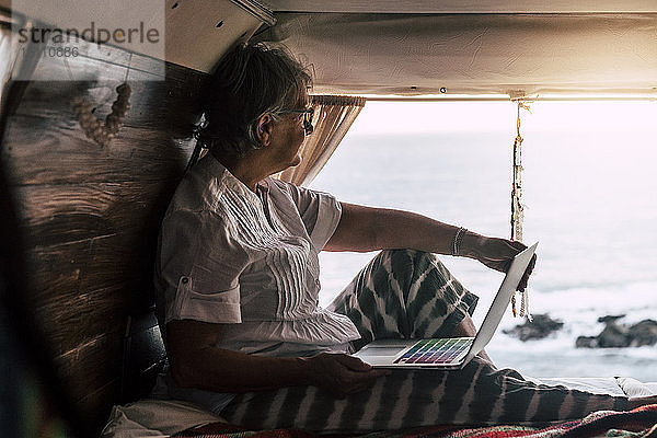 Ältere Frau sitzt im Oldtimer-Van am Meer und benutzt einen Laptop