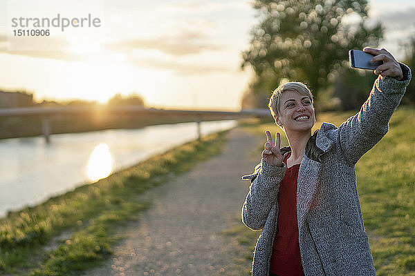 Porträt einer grinsenden Frau  die sich bei Sonnenuntergang mit einem Smartphone selbstständig macht