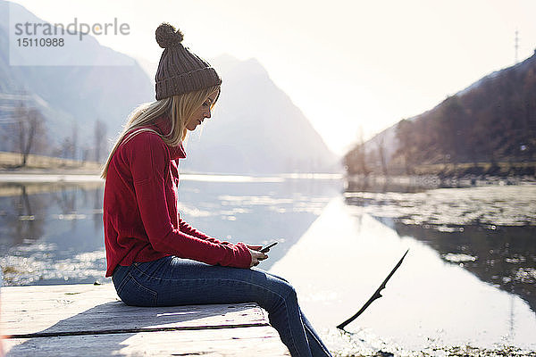 Junge blonde Frau sitzt im Winter auf einem Steg an einem See