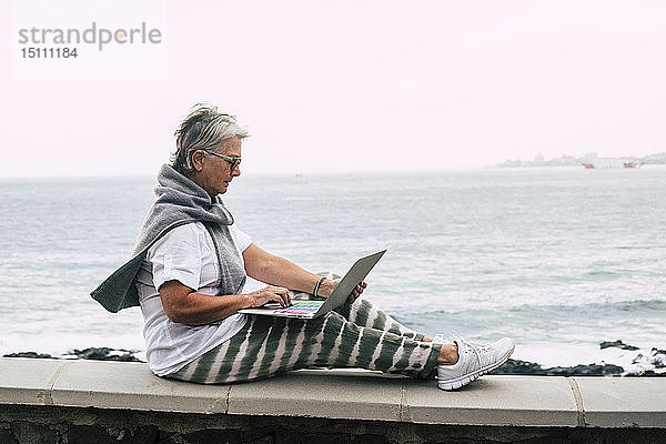 Ältere Frau sitzt auf einer Mauer am Meer und benutzt einen Laptop