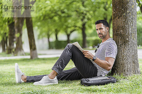 Mann lehnt an einem Baum im Park und liest Zeitung