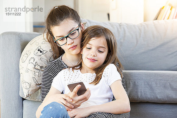 Zwei Schwestern sitzen vor der Couch und schauen auf das Smartphone