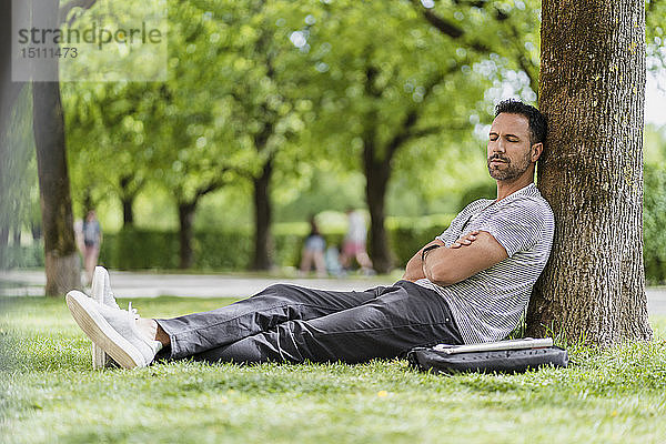 Mann lehnt an einem Baum im Park und hält ein Nickerchen