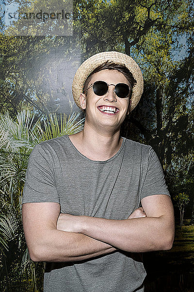 Junger Mann mit Hut und Sonnenbrille  vor Fototapete stehend  mit verschränkten Armen