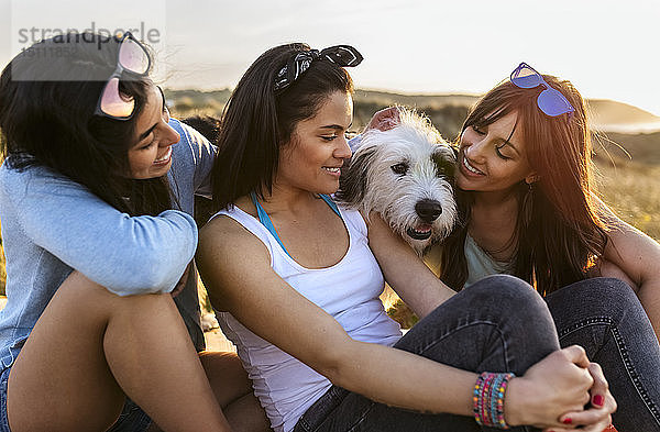 Drei glückliche Frauen mit Hund sitzen bei Sonnenuntergang in den Dünen