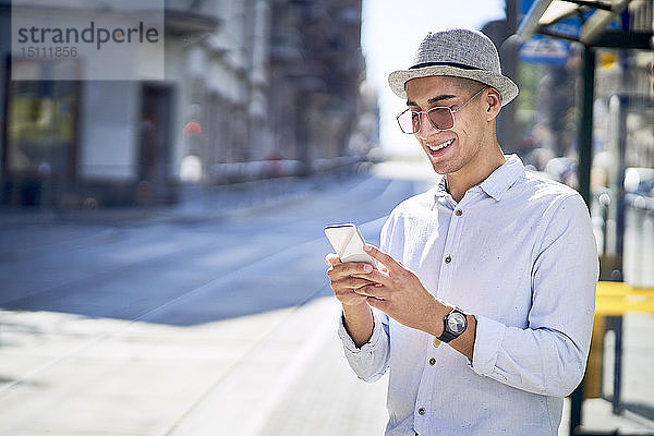 Glücklicher junger Mann überprüft sein Telefon an einer Straßenbahnhaltestelle in der Stadt