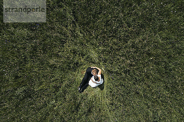 Luftaufnahme einer schwangeren Braut mit ihrem Mann auf einer Wiese sitzend