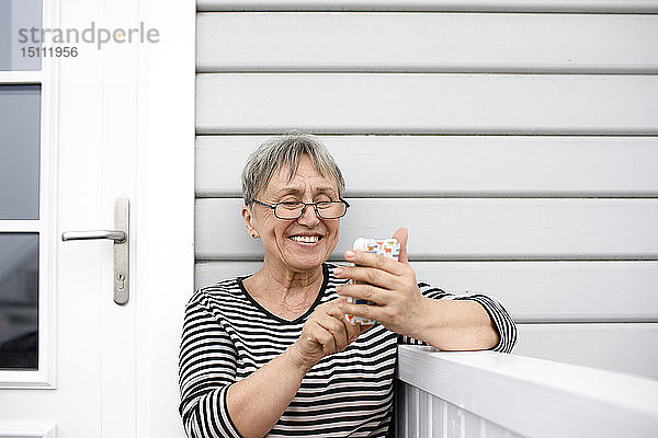 Glückliche ältere Frau benutzt Handy auf der Veranda ihres Hauses