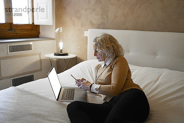 Reife Geschäftsfrau  die mit einem Laptop auf dem Bett liegt und ein Mobiltelefon benutzt