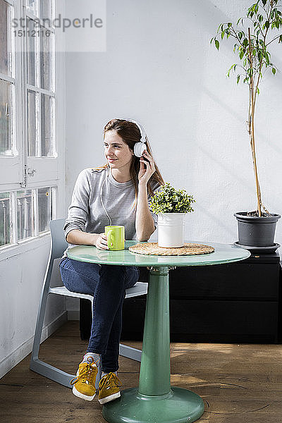Junge Frau  die zu Hause mit Kaffeetasse und Kopfhörern am Fenster sitzt