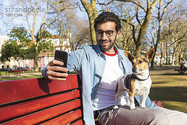 Porträt eines jungen Mannes  der mit seinem Hund auf einer Parkbank sitzt und sich mit seinem Smartphone selbstständig macht