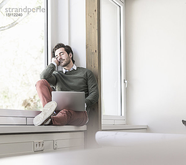 Junger Geschäftsmann mit Laptop sitzt auf der Fensterbank und macht eine Pause