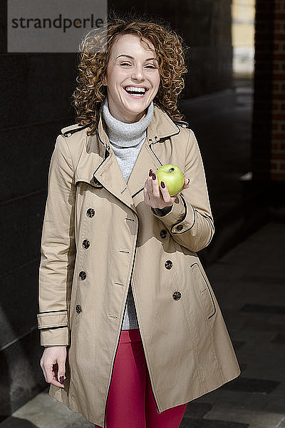 Bildnis einer lachenden Frau mit grünem Apfel in beigem Trenchcoat und Rollkragenpullover