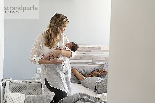 Mutter hält ihr Neugeborenes im Schlafzimmer  während der Vater im Hintergrund schläft