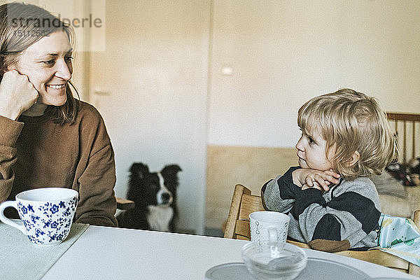 Lächelnde Mutter und Tochter sitzen zu Hause am Tisch mit Hund im Hintergrund