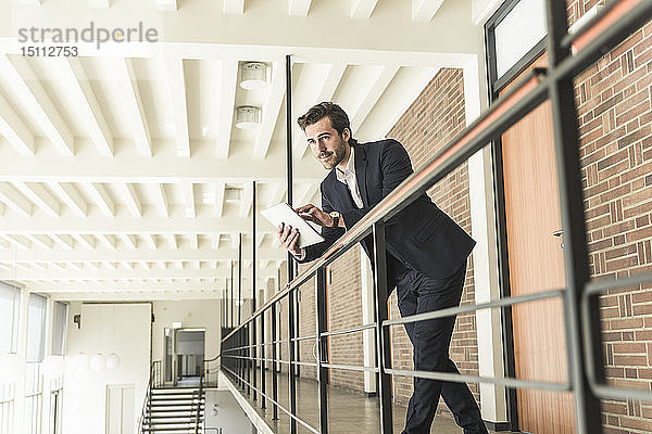 Junger Geschäftsmann steht auf Galerie in modernem Bürogebäude und benutzt digitales Tablet