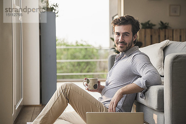 Junger Mann sitzt zu Hause auf dem Boden  benutzt Laptop  trinkt Kaffee