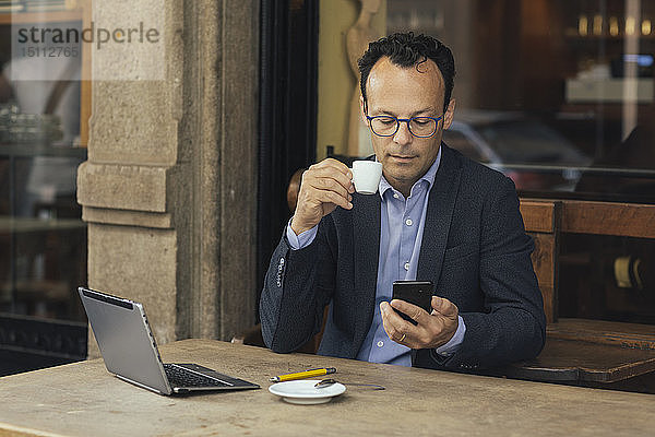 Geschäftsmann mit Laptop in einem Café  der auf sein Smartphone schaut