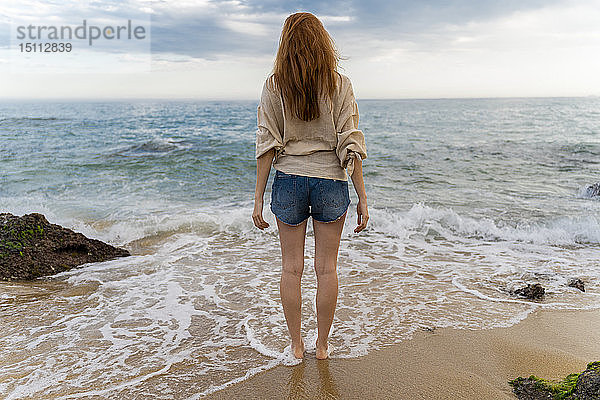 Rückenansicht einer rothaarigen jungen Frau  die am Strand steht und aufs Meer blickt