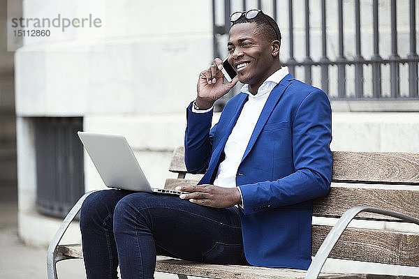 Junger Geschäftsmann in blauer Anzugjacke sitzt auf Bank und benutzt Smartphone
