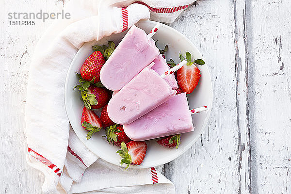 Hausgemachte Erdbeer-Joghurt-Eislollys mit frischen Erdbeeren in einer Schale auf weißem Holz